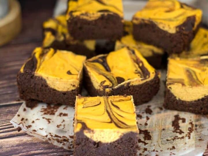 A batch of chocolate and pumpkin cheesecake swirl brownies arranged on a parchment paper atop a wooden surface.