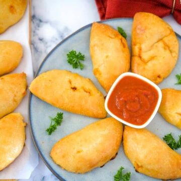 A plate of low carb turkey calzones arranged in a circle around a small bowl of red dipping sauce, garnished with parsley, with more empanadas on the side.