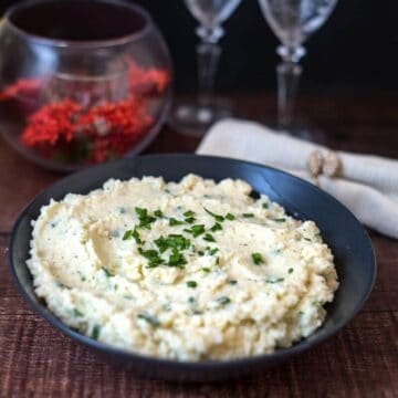 A bowl of keto cauliflower mash garnished with chopped herbs, placed on a wooden table with a folded napkin and wine glasses in the background.