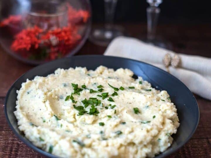 A bowl of keto cauliflower mash garnished with chopped herbs, placed on a wooden table with a folded napkin and wine glasses in the background.