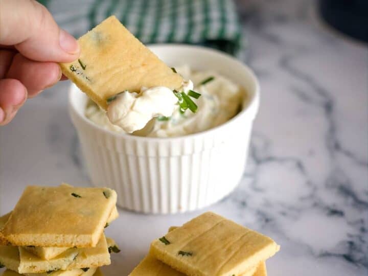 A hand dips a square cracker with green bits into a white bowl of creamy sour cream and chive dip, with more crackers stacked beside it on a marble surface. A green and white cloth is in the background.