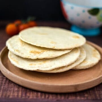 A stack of four keto almond flour tortillas on a round wooden plate with tomatoes and a bowl in the background.