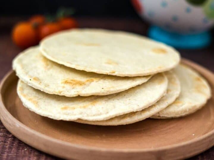 A stack of four keto almond flour tortillas on a round wooden plate with tomatoes and a bowl in the background.