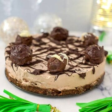 A chocolate mousse cake topped with chocolate truffles and drizzled with dark chocolate, set on a crust, with green paper decorations and a lit background, reminiscent of a no bake Irish cream cheesecake.