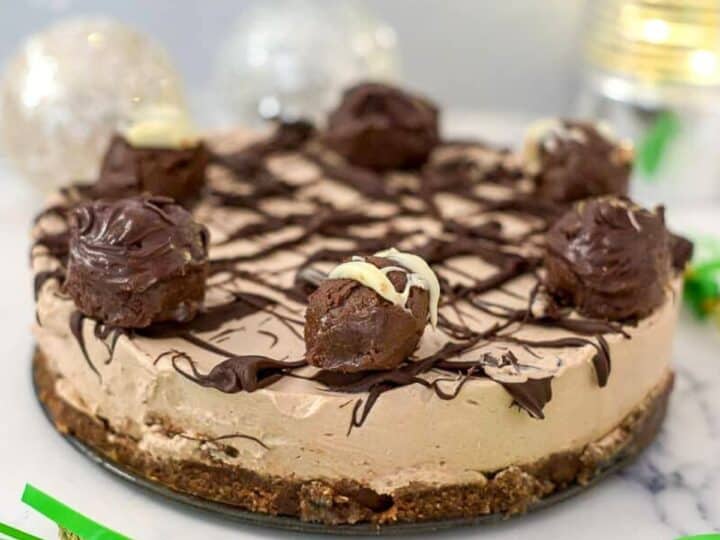 A chocolate mousse cake topped with chocolate truffles and drizzled with dark chocolate, set on a crust, with green paper decorations and a lit background, reminiscent of a no bake Irish cream cheesecake.