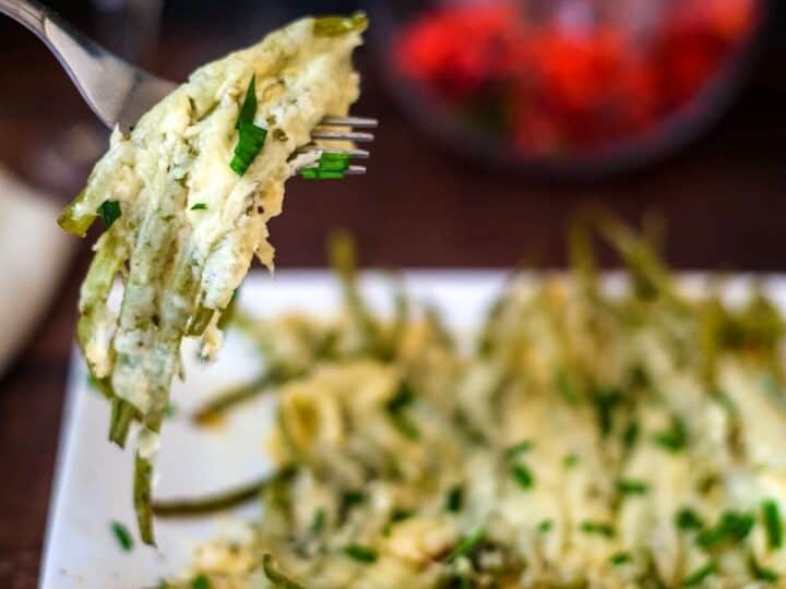 A fork lifts a portion of cheesy keto green bean casserole from a white plate.