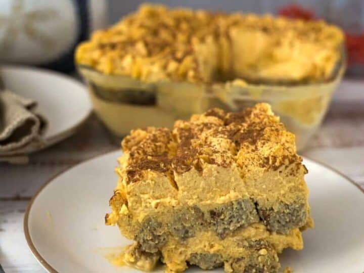 A plate containing a slice of layered pumpkin tiramisu with crumbled topping, in front of a glass dish with more of the same dessert.