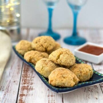 A blue rectangular plate holds several breaded cauliflower balls with a small square dish of sauce. Two filled wine glasses and a napkin are in the background on a rustic wooden surface.