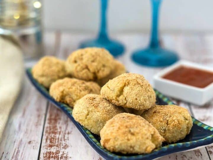 A blue rectangular plate holds several breaded cauliflower balls with a small square dish of sauce. Two filled wine glasses and a napkin are in the background on a rustic wooden surface.