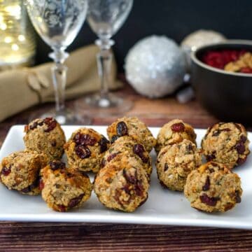 A white plate holding multiple round oat and cranberry cookies on a wooden table. In the background, two wine glasses, a napkin, and a bowl of cranberry stuffing balls complete the cozy scene.