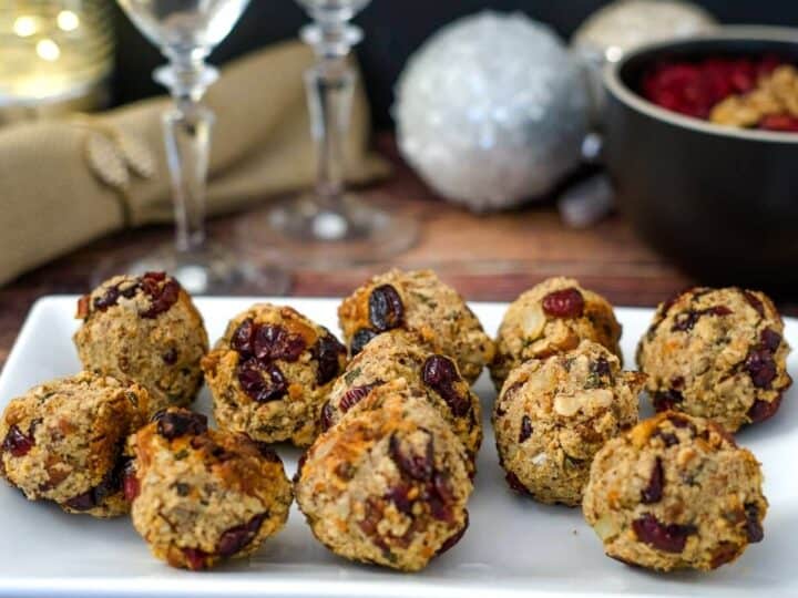 A white plate holding multiple round oat and cranberry cookies on a wooden table. In the background, two wine glasses, a napkin, and a bowl of cranberry stuffing balls complete the cozy scene.