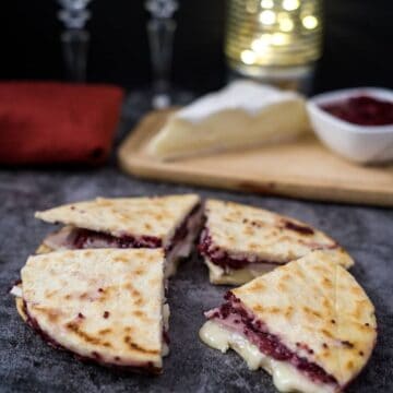 A sliced low carb quesadilla with melted cheese and cranberry sauce, placed on a dark surface. In the background are a block of cheese, a bowl of cranberry sauce, a red cloth, and blurred lights in a jar.