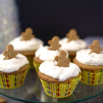 Keto gingerbread cupcakes with white frosting topped with small gingerbread man cookies are displayed on a glass stand. The cupcakes are in yellow polka-dotted wrappers.