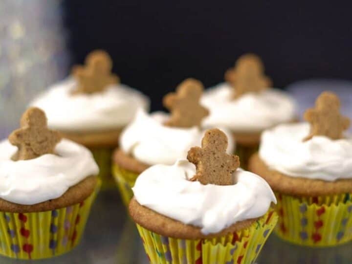 Keto gingerbread cupcakes with white frosting topped with small gingerbread man cookies are displayed on a glass stand. The cupcakes are in yellow polka-dotted wrappers.