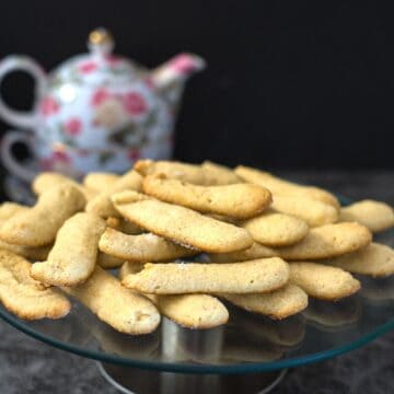 A glass plate filled with keto lady fingers is placed on a dark surface. A floral teapot is in the background.