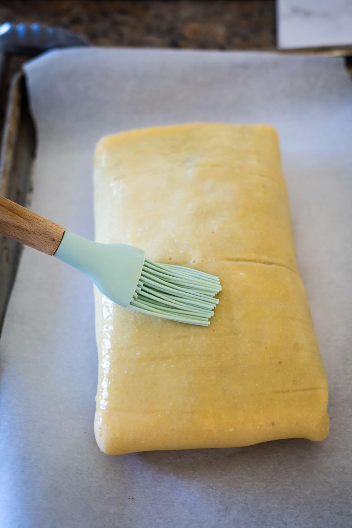 A person is using a brush to brush a piece of dough onto a baking sheet to prepare a vegetable wellington.