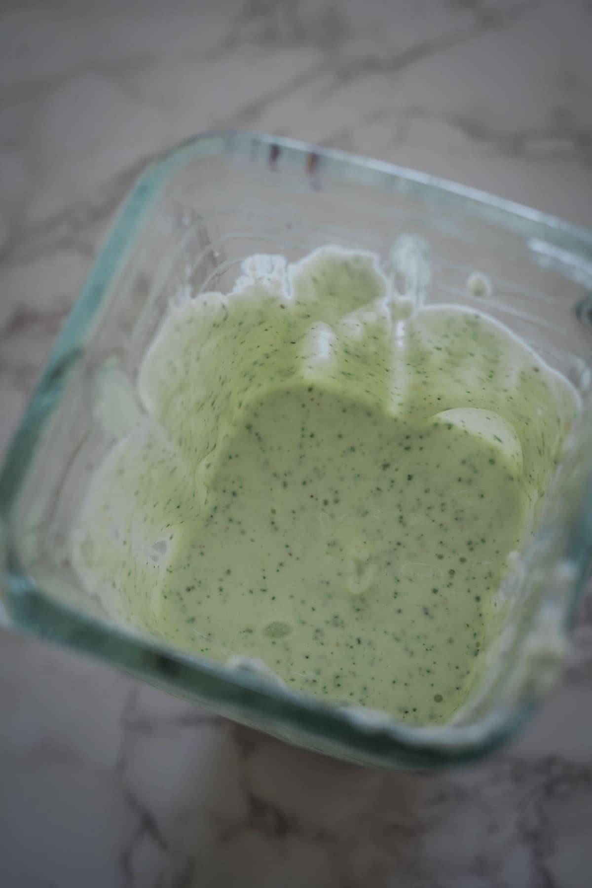 A glass container with a creamy green sauce or dip, speckled with small herb bits, on a marble surface.