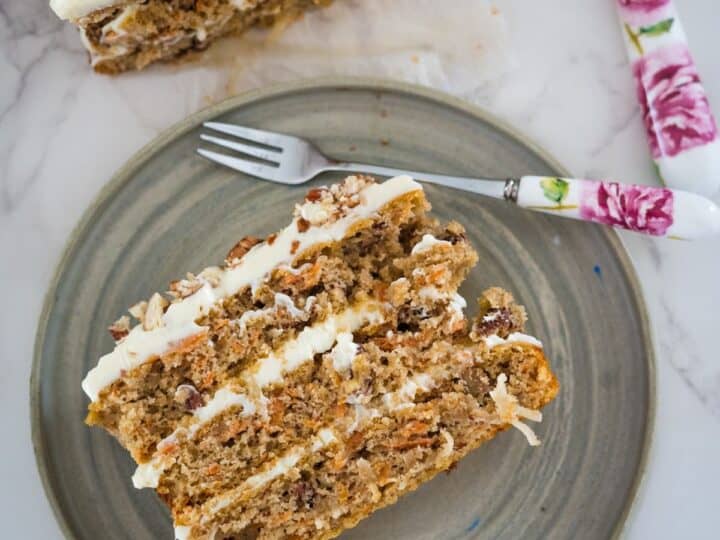 A slice of carrot cake with cream cheese frosting on a ceramic plate next to a floral-patterned fork.