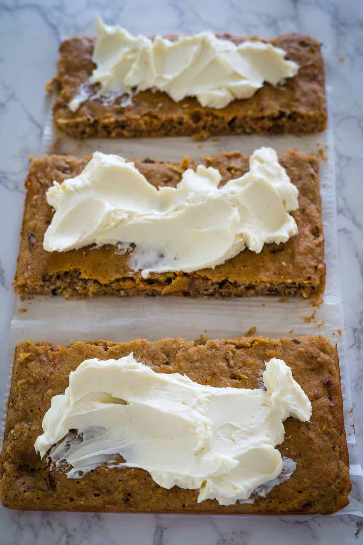 Three keto carrot cake bars with cream cheese frosting on a white marble surface.