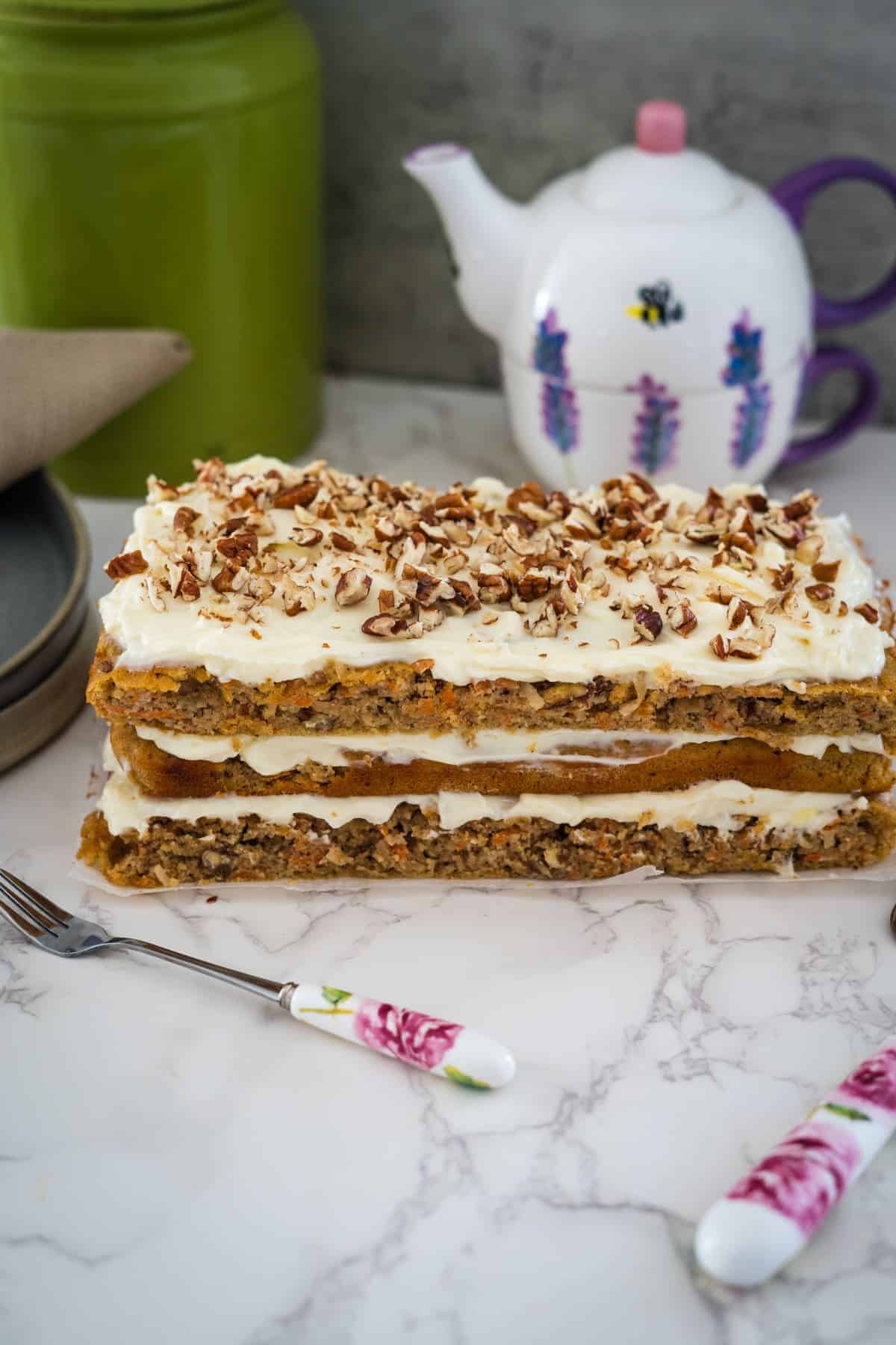 A layered carrot cake with cream cheese frosting and chopped nuts, served with a teapot on a marble surface.