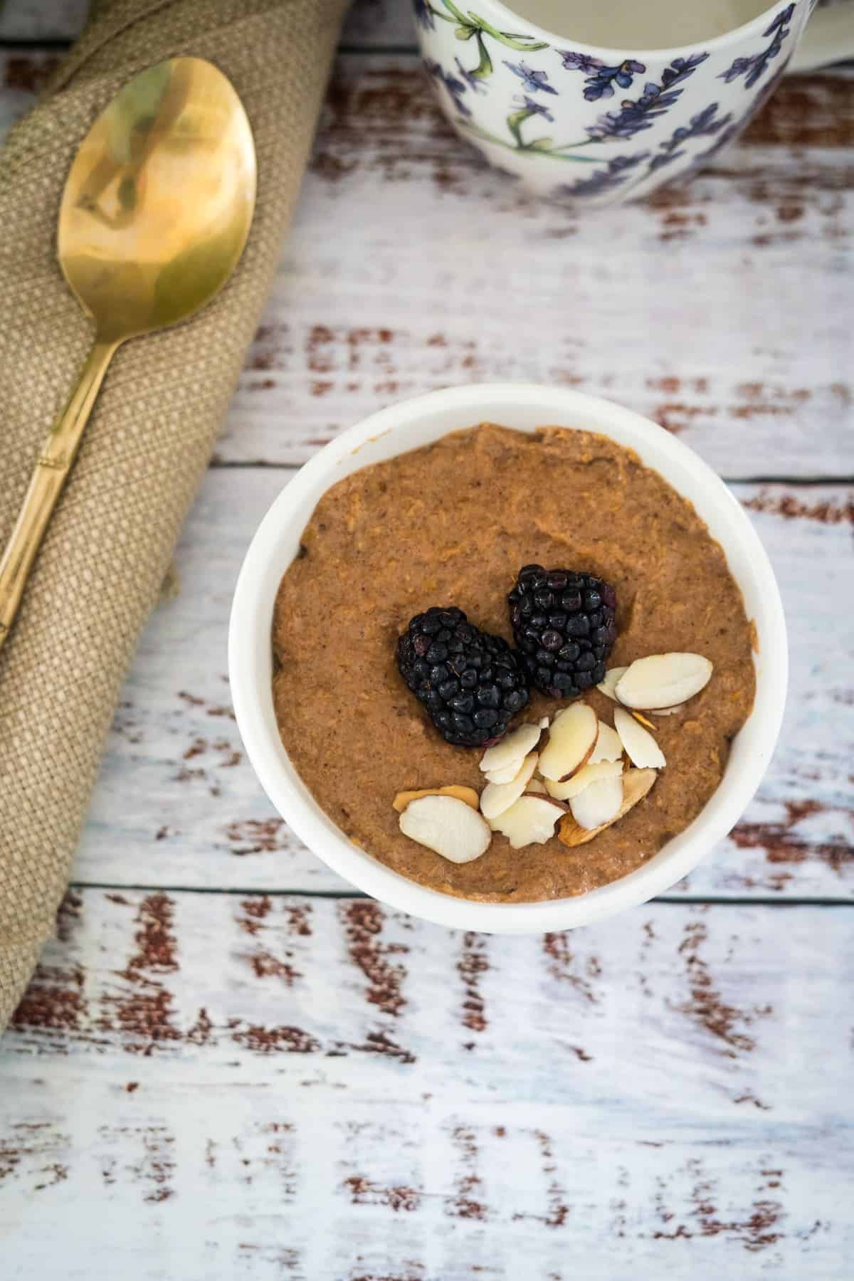 A bowl of oatmeal garnished with almonds and blackberries, with a spoon and a cup in the background, on a rustic wooden surface.