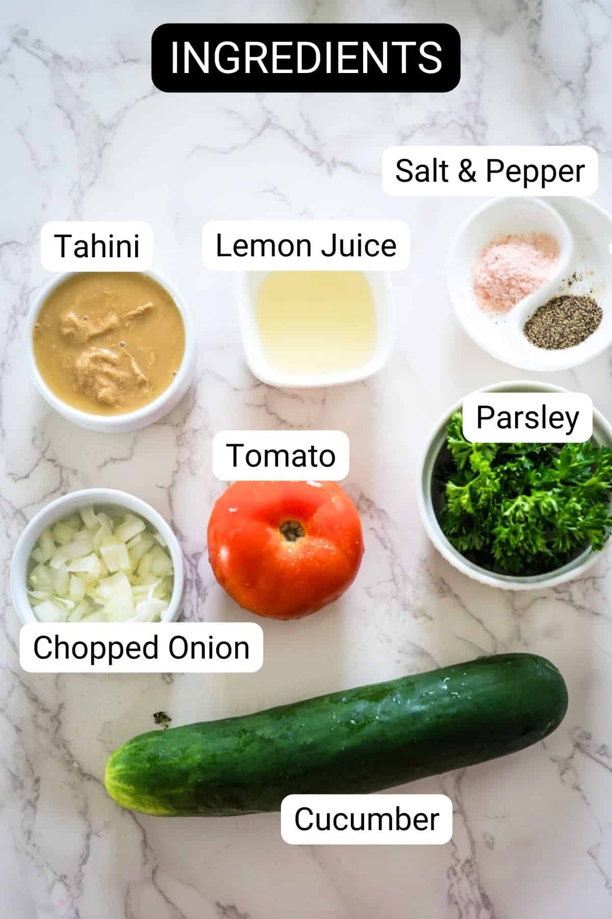 An assortment of fresh ingredients labeled for a recipe, including tahini, lemon juice, salt & pepper, chopped onion, tomato, parsley, and cucumber, arranged on a marble surface.