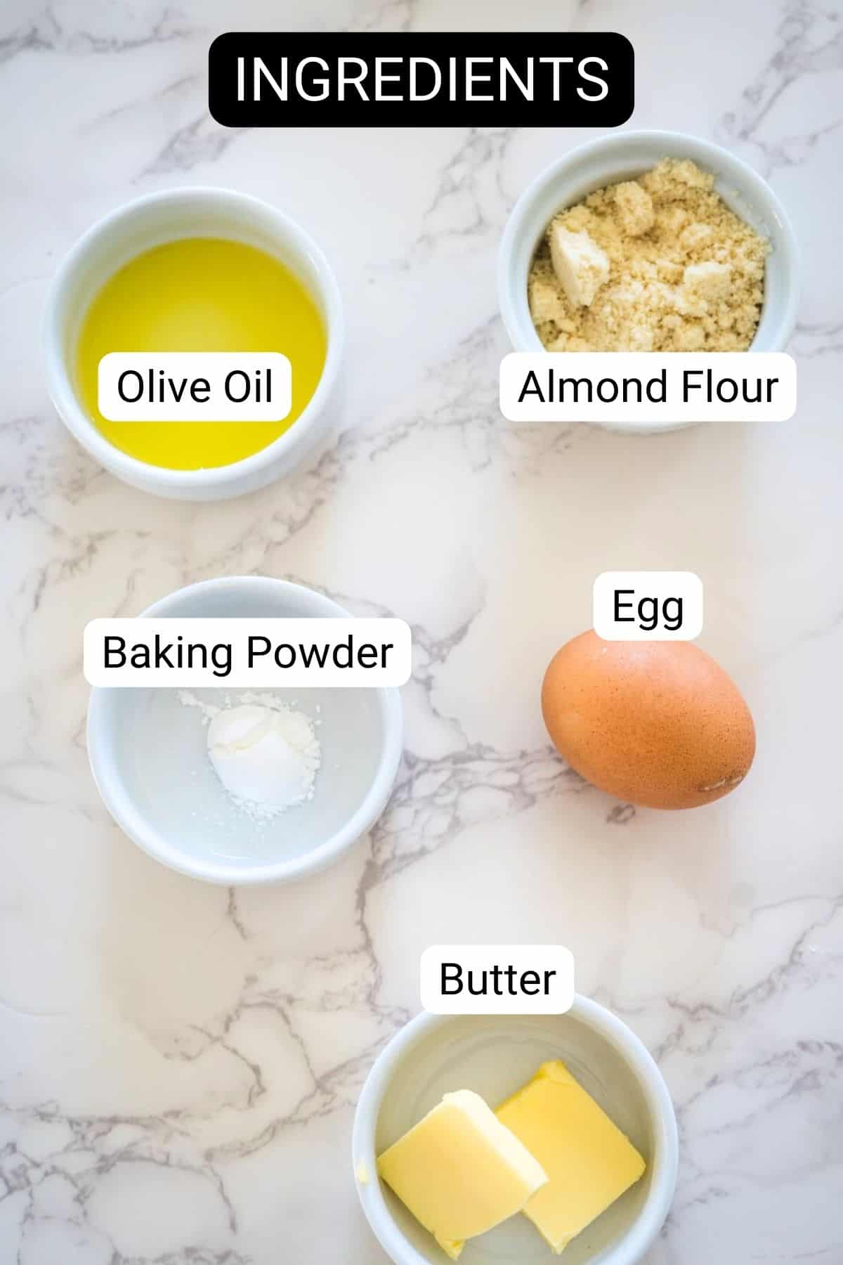 Various baking ingredients labeled on a marble surface, including olive oil, almond flour, keto bread crumbs, an egg, and butter.