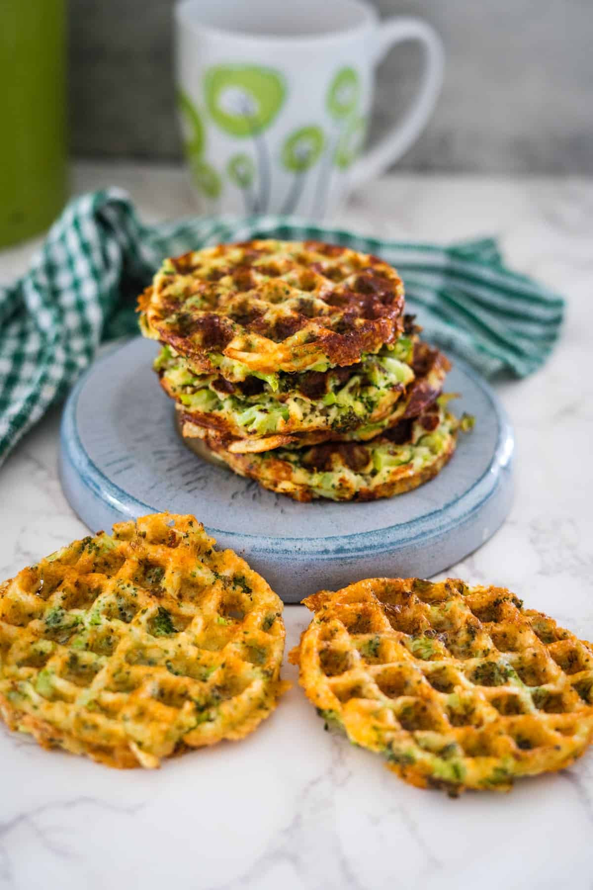 A stack of crispy zucchini fritters served on a blue plate with a mug and a green checked napkin in the background.