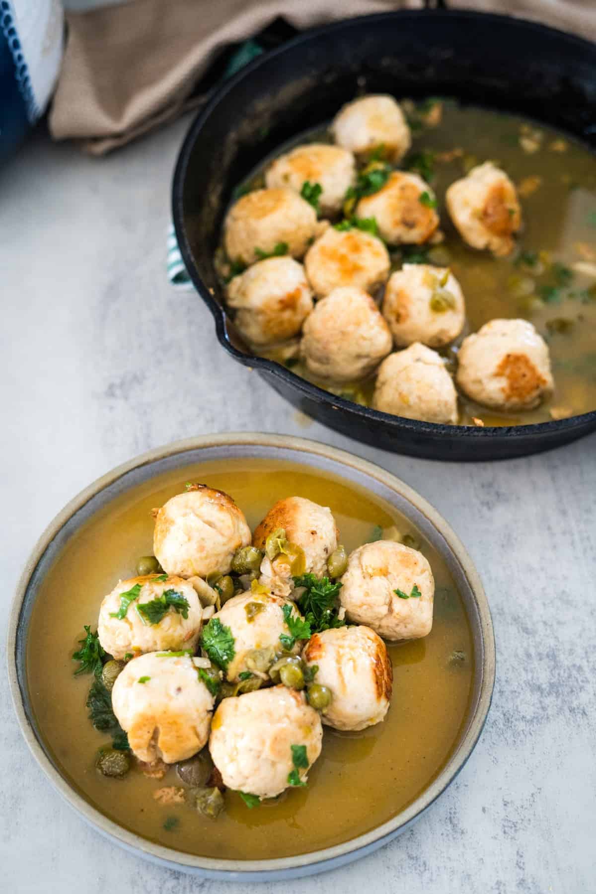 A bowl of chicken meatballs with peas and parsley in broth, alongside a cast iron skillet containing more servings.