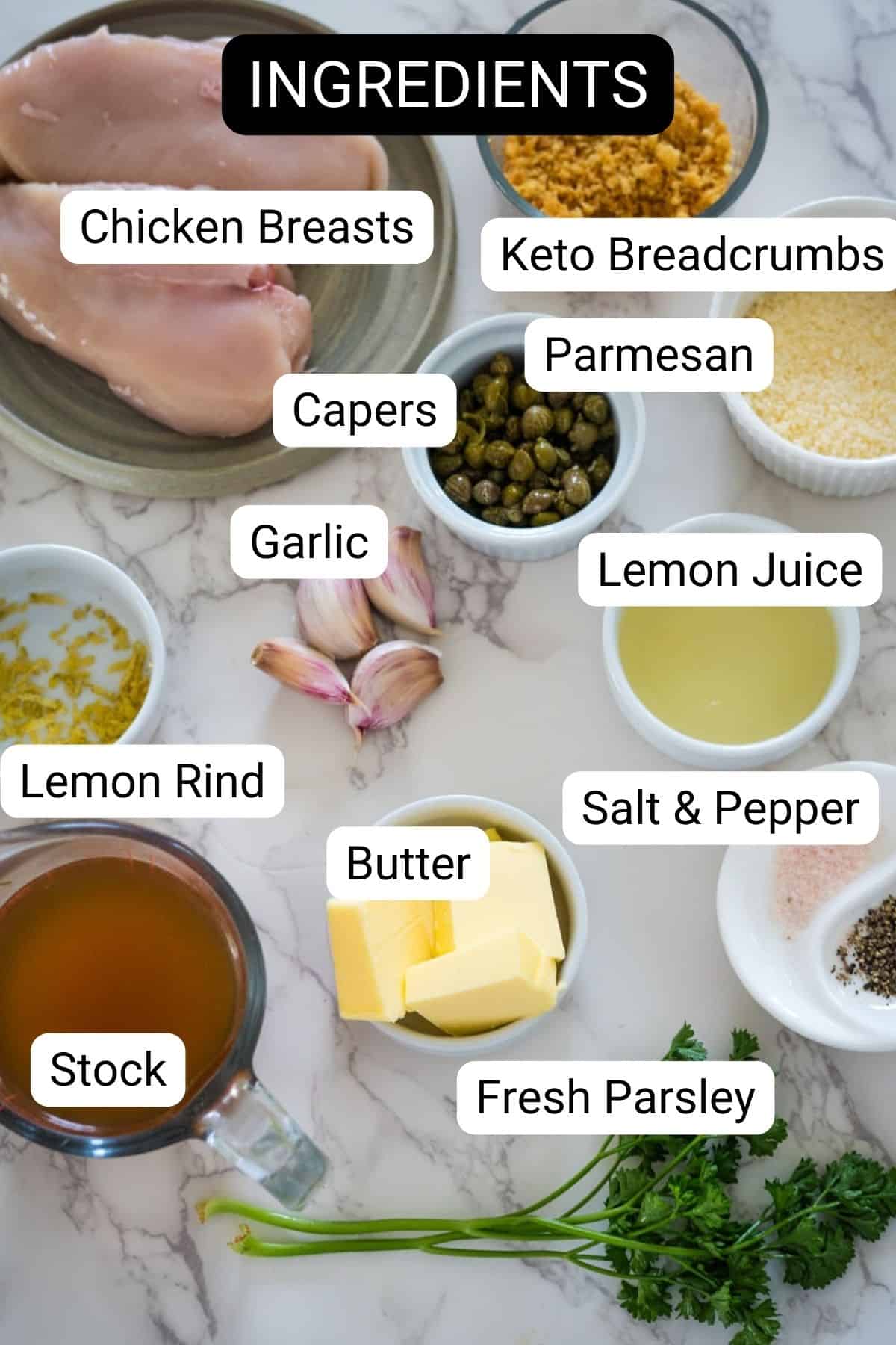 Various ingredients for a recipe laid out on a countertop, including chicken, breadcrumbs, capers, garlic, lemon juice, butter, and fresh parsley, labeled for clarity.