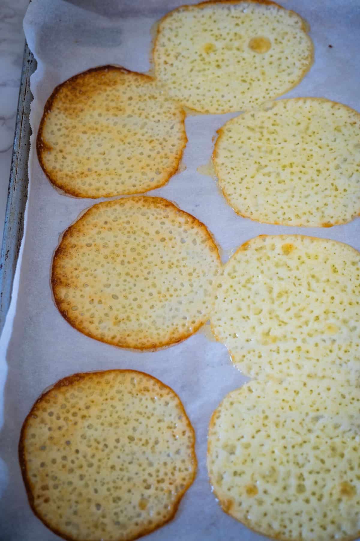 Thin, crispy cheese crisps cooling on parchment paper with a light golden color and visible porous texture.