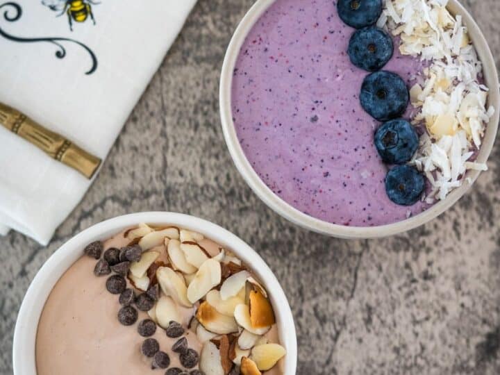 Two bowls of smoothie topped with blueberries, coconut, almonds, and chocolate chips, served on a concrete surface next to a napkin with a bee design.