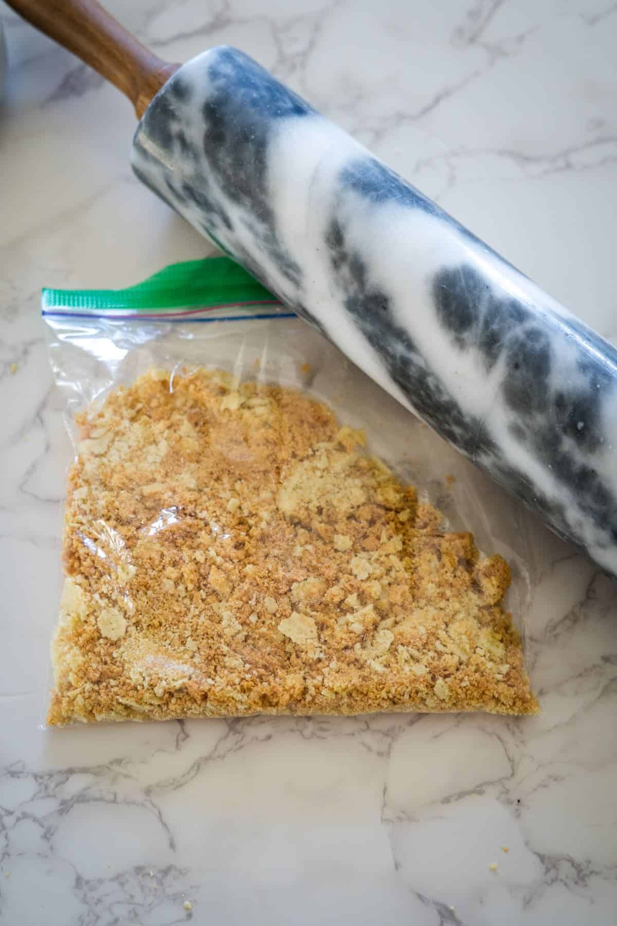 A marbled rolling pin on a marble countertop next to a plastic bag filled with crushed crackers.