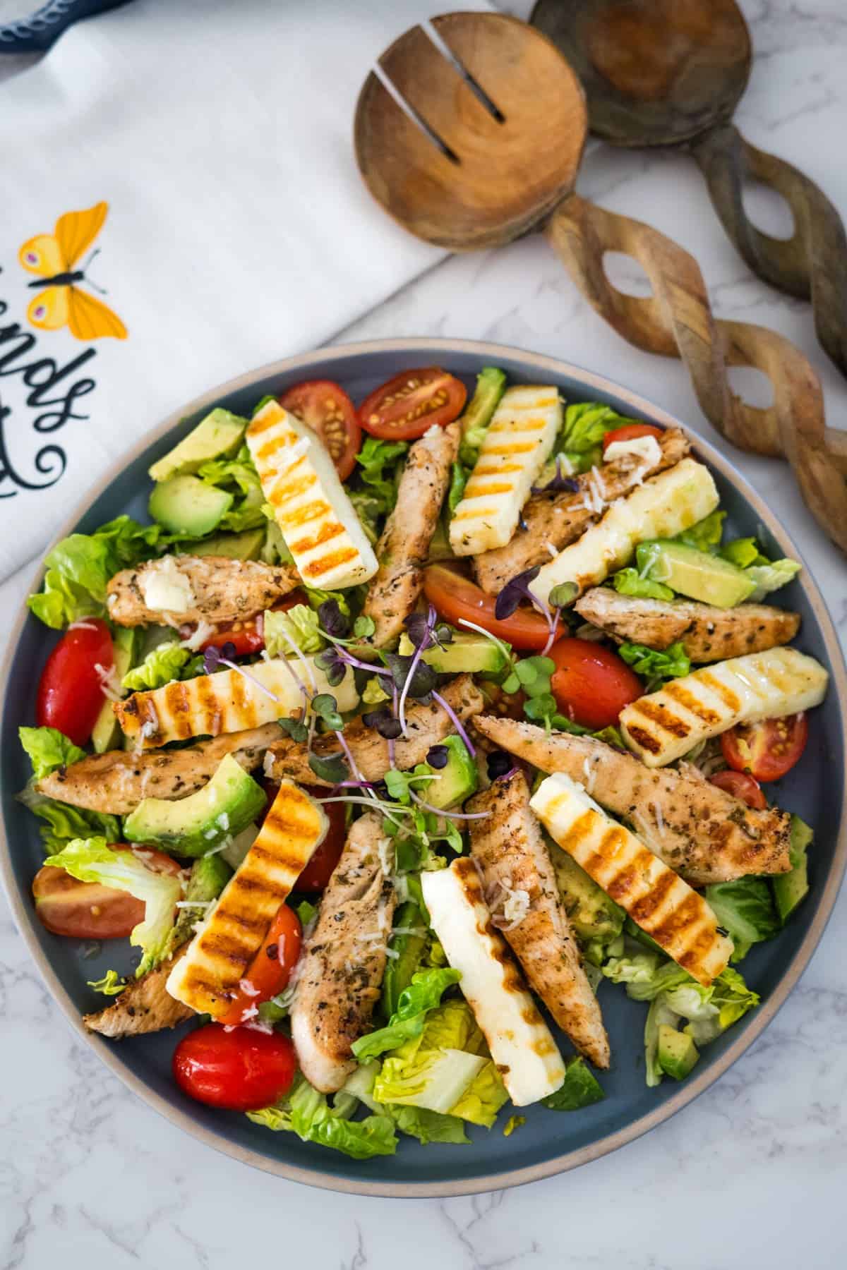 A fresh salad with grilled chicken strips, avocado slices, cherry tomatoes, and halloumi cheese, garnished with microgreens on a gray plate.