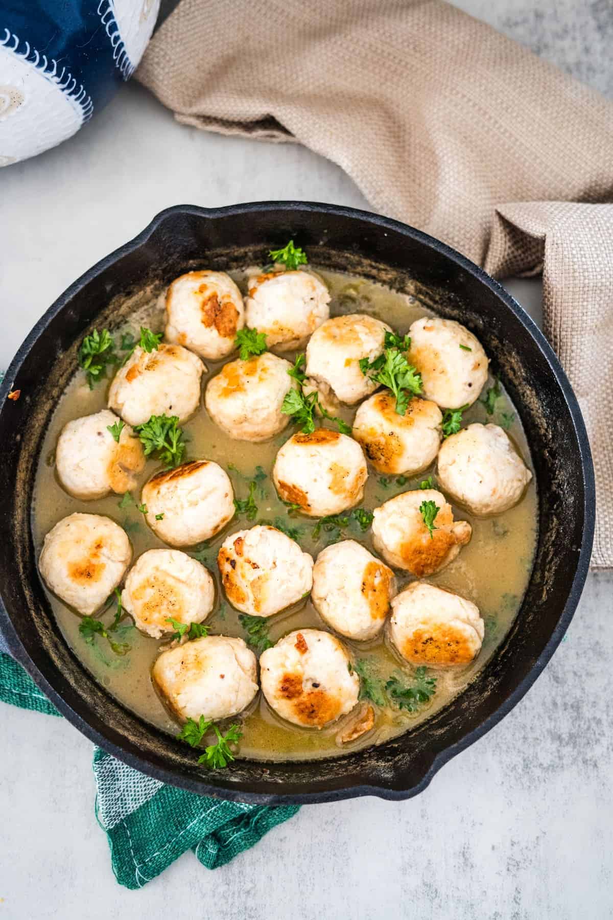 A cast iron skillet filled with seared scallops garnished with parsley, accompanied by a beige napkin on a light surface.