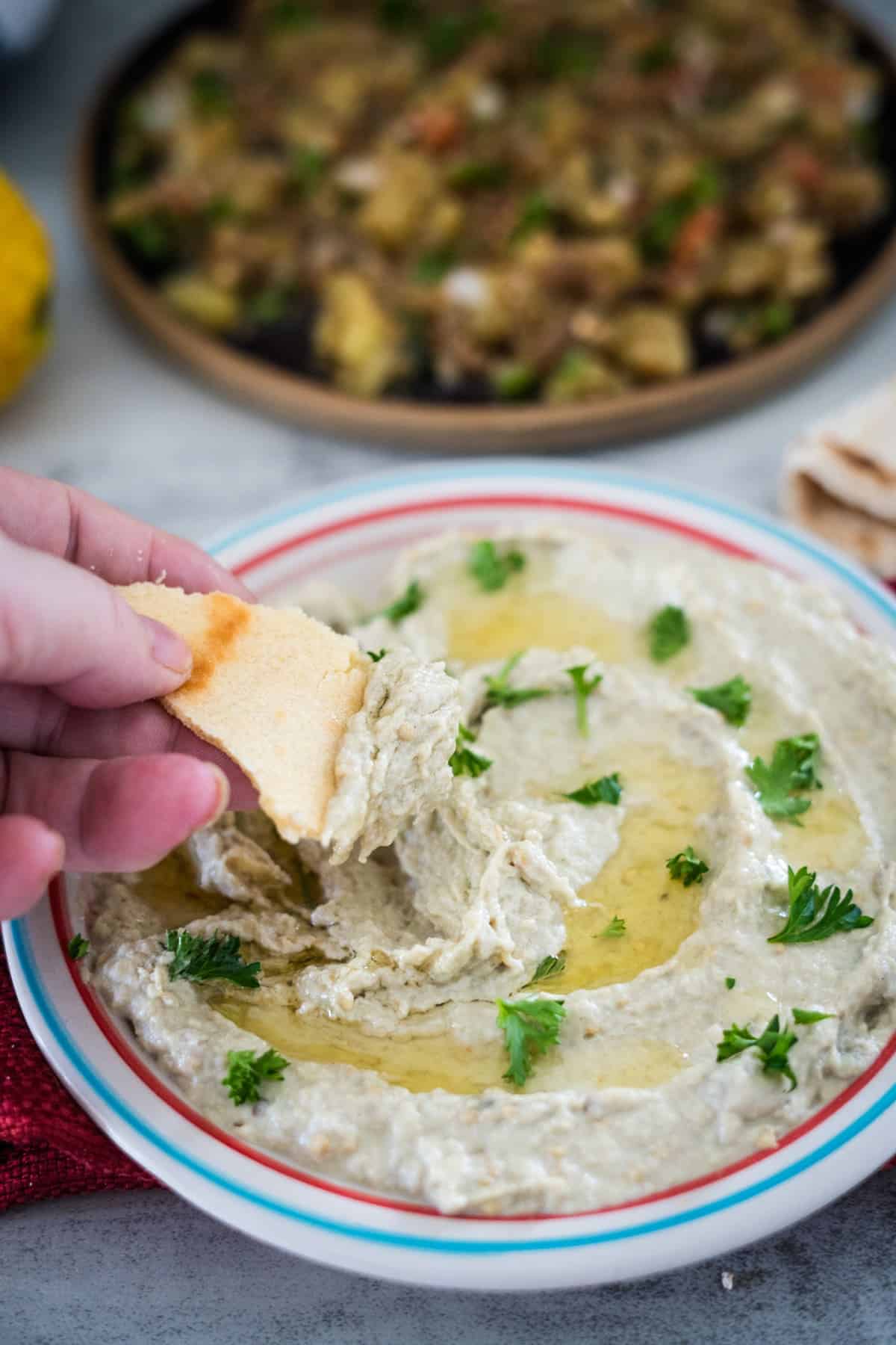 Hand dipping pita bread into a bowl of hummus garnished with olive oil and parsley.