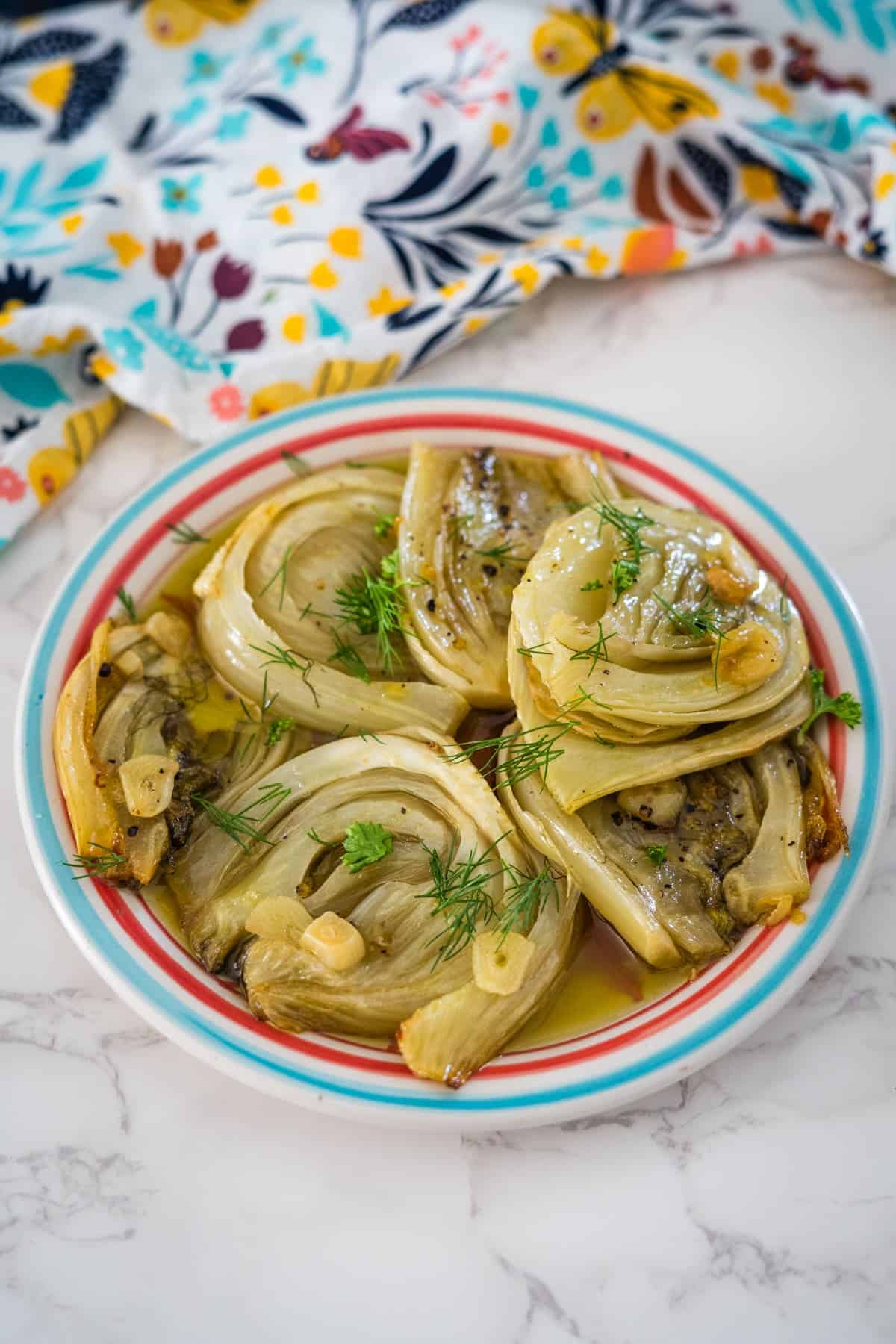 A plate of roasted fennel confit garnished with dill on a striped plate, set on a marble surface with a colorful floral cloth.