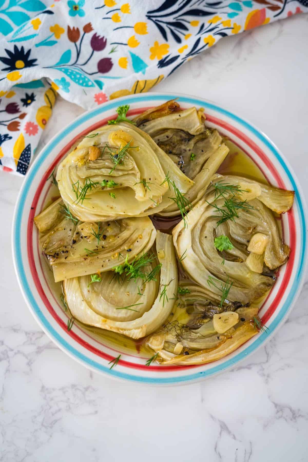 A plate of braised fennel halves garnished with herbs, pictured on a marble countertop next to a colorful cloth.