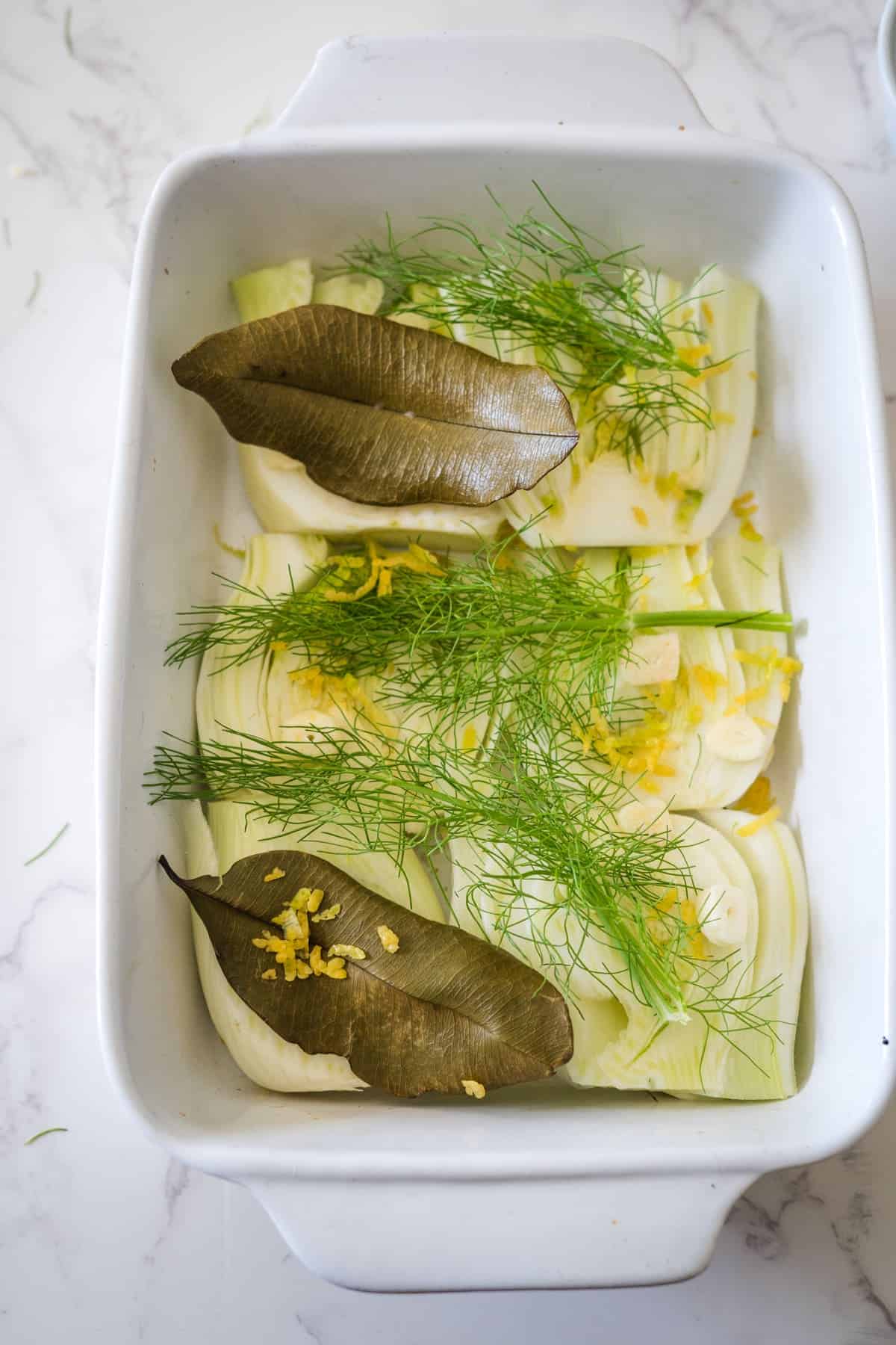 Baking dish containing fennel confit and leeks topped with fresh dill and bay leaves, ready for cooking.