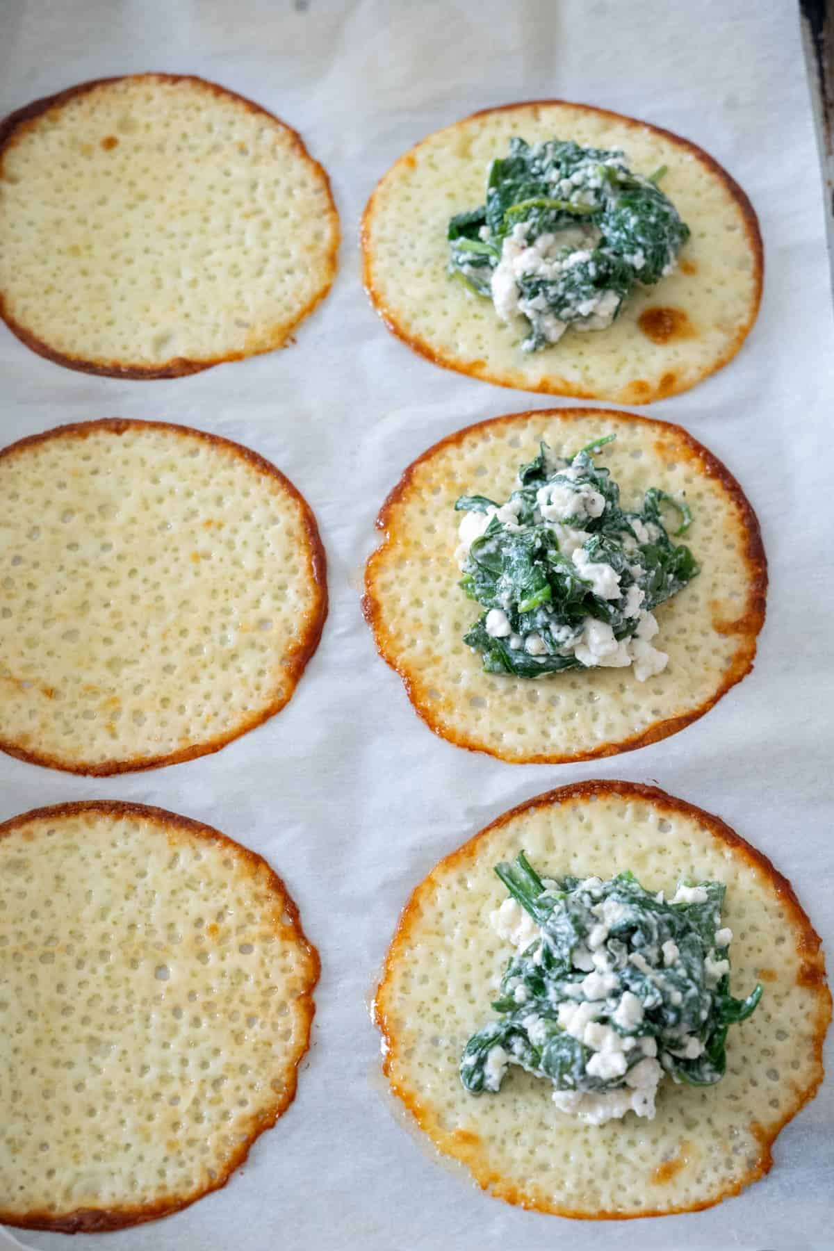 Six golden-brown cheese crisps on parchment paper, three topped with a spinach and cheese mixture.