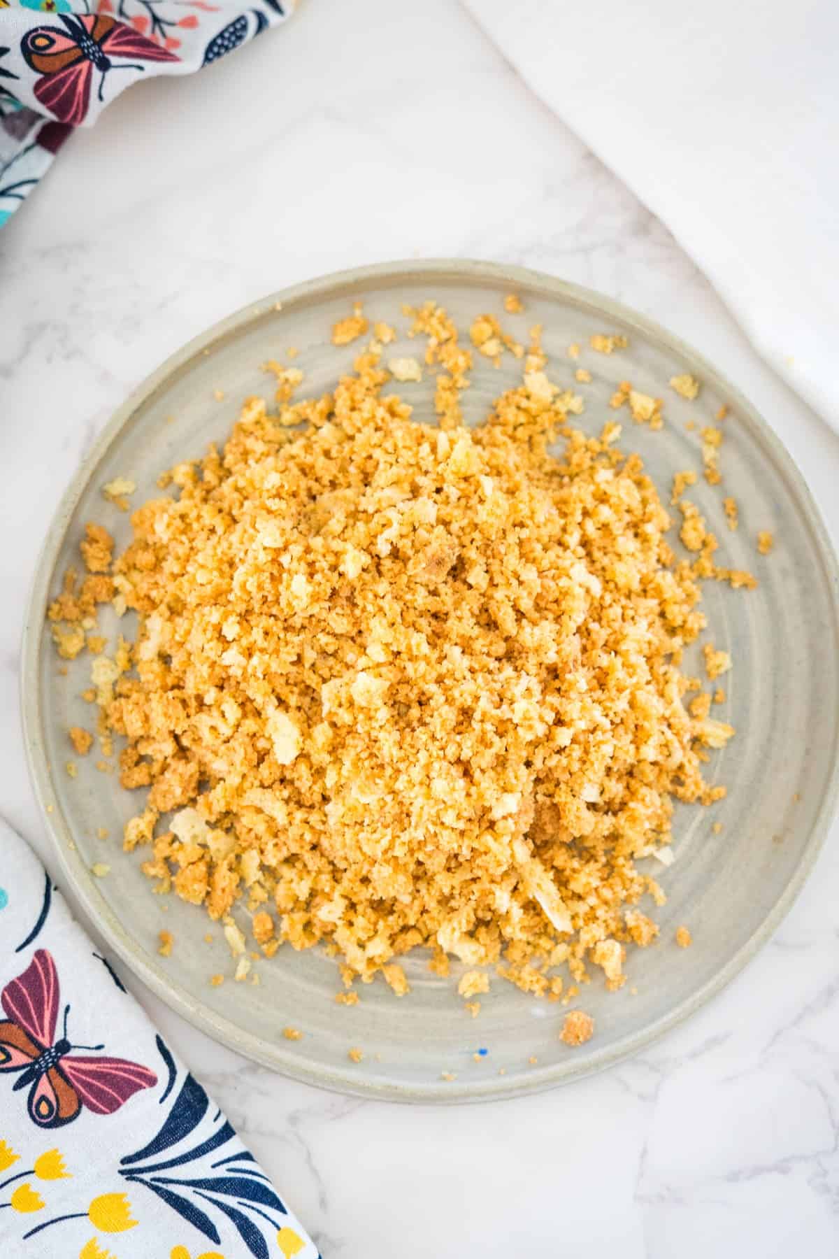 A bowl of keto bread crumbs on a light ceramic plate, placed on a white surface next to a floral fabric.