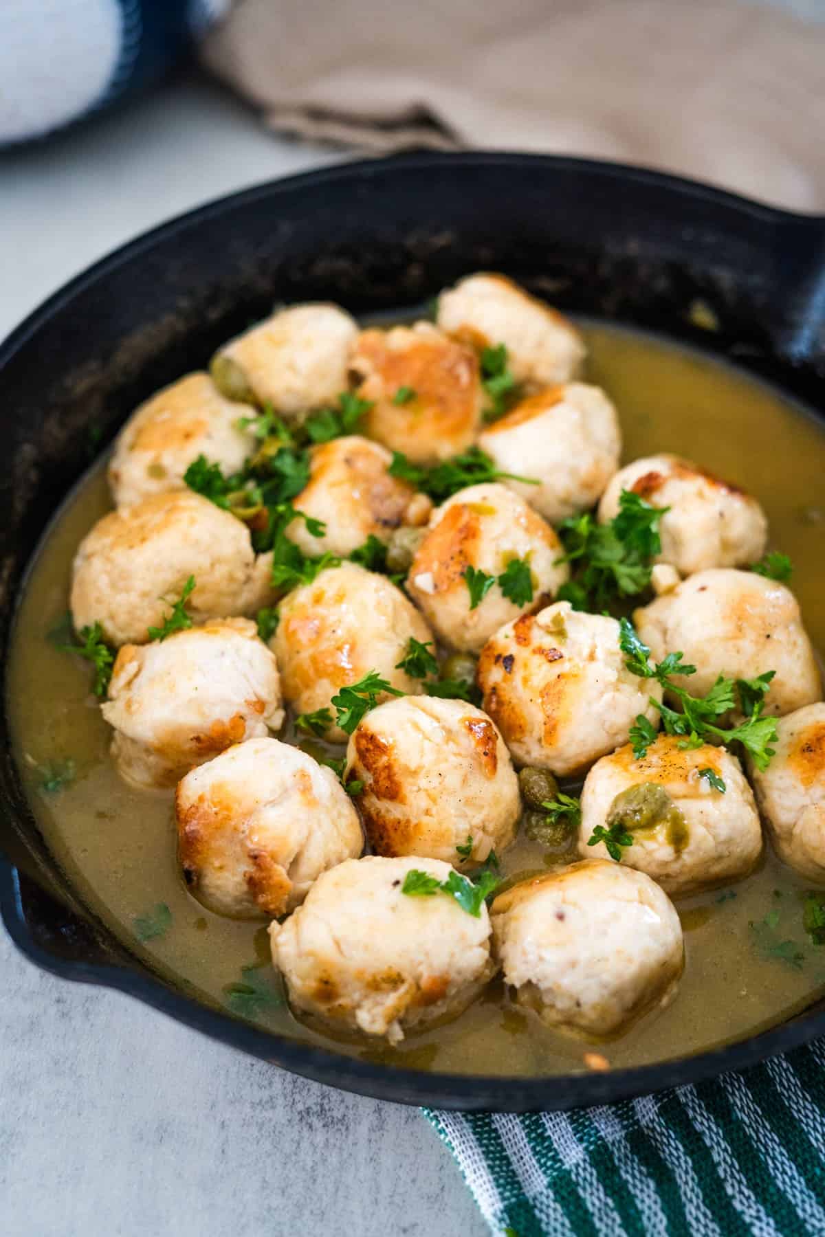 A skillet of chicken meatballs in a savory sauce, garnished with chopped parsley, served on a striped cloth.