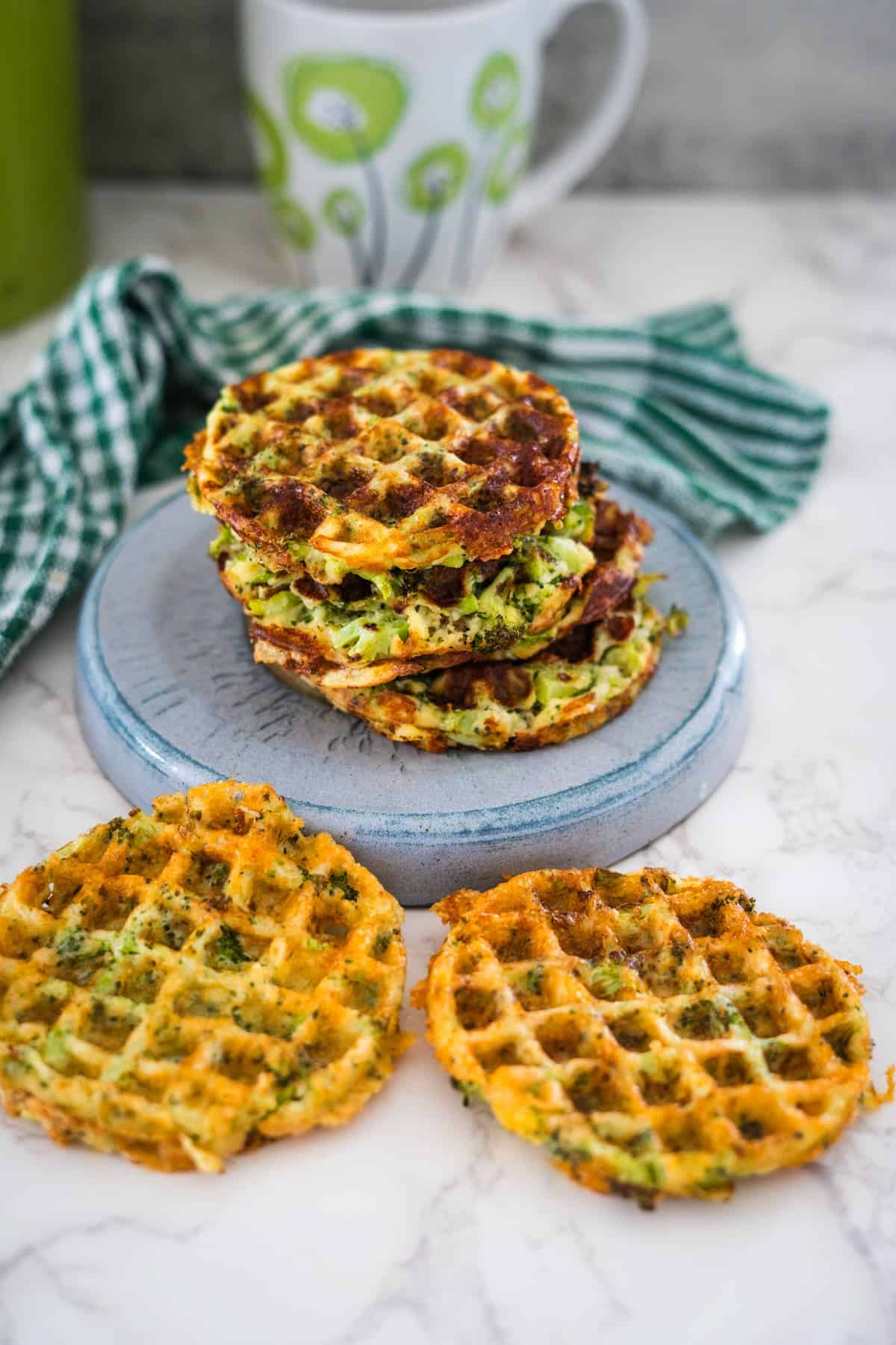 Three broccoli cheddar waffles stacked on a blue plate with a mug and green-striped napkin in the background.