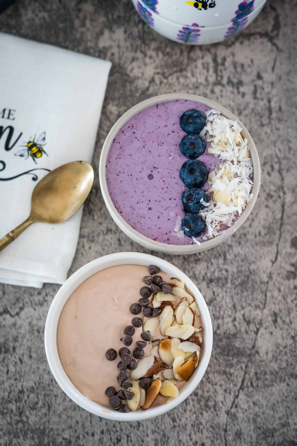 Two bowls of smoothie bowls on a textured surface, one topped with blueberries and coconut, the other with almond slices and chocolate chips, next to a patterned cup and a napkin filled with