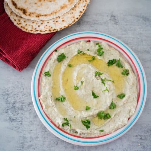 A bowl of hummus garnished with olive oil and parsley, served with flatbread on the side.