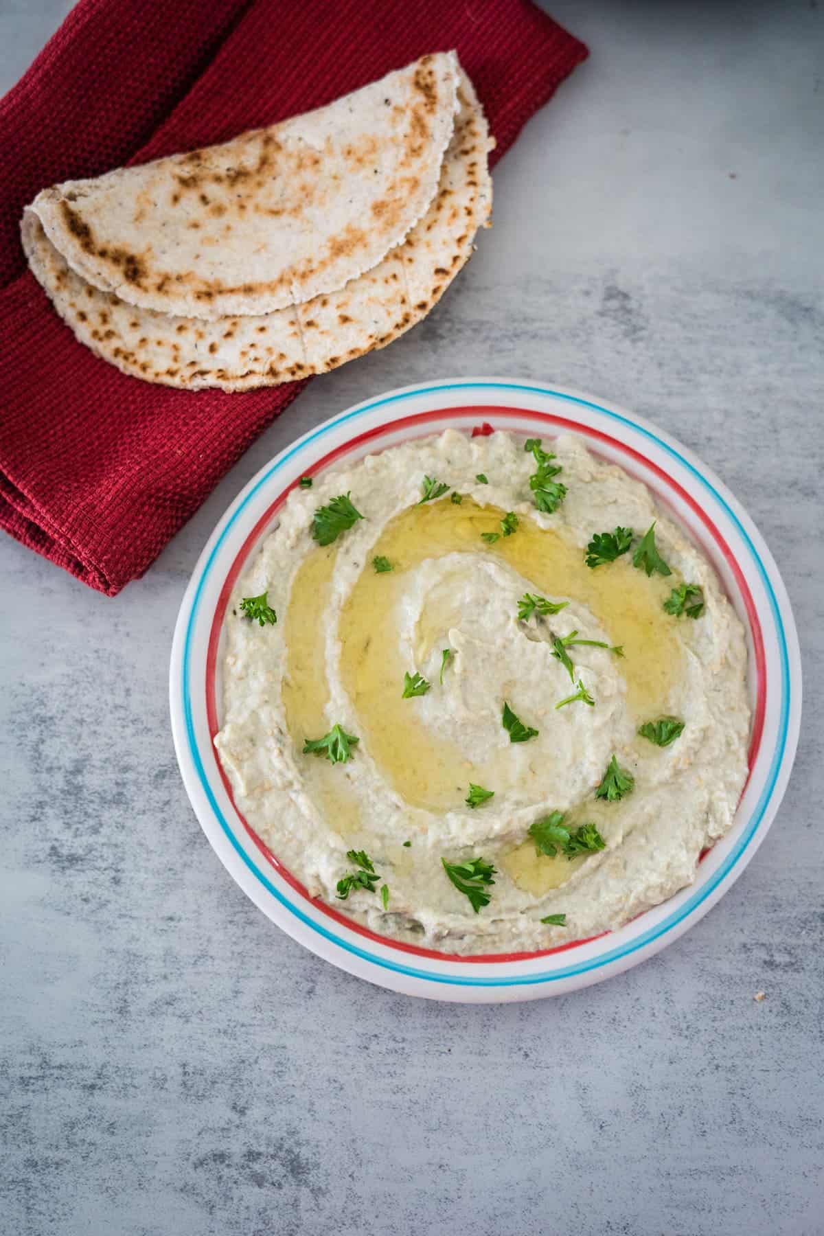 Bowl of mutabal garnished with parsley and olive oil, served with pieces of pita bread on a marble surface.