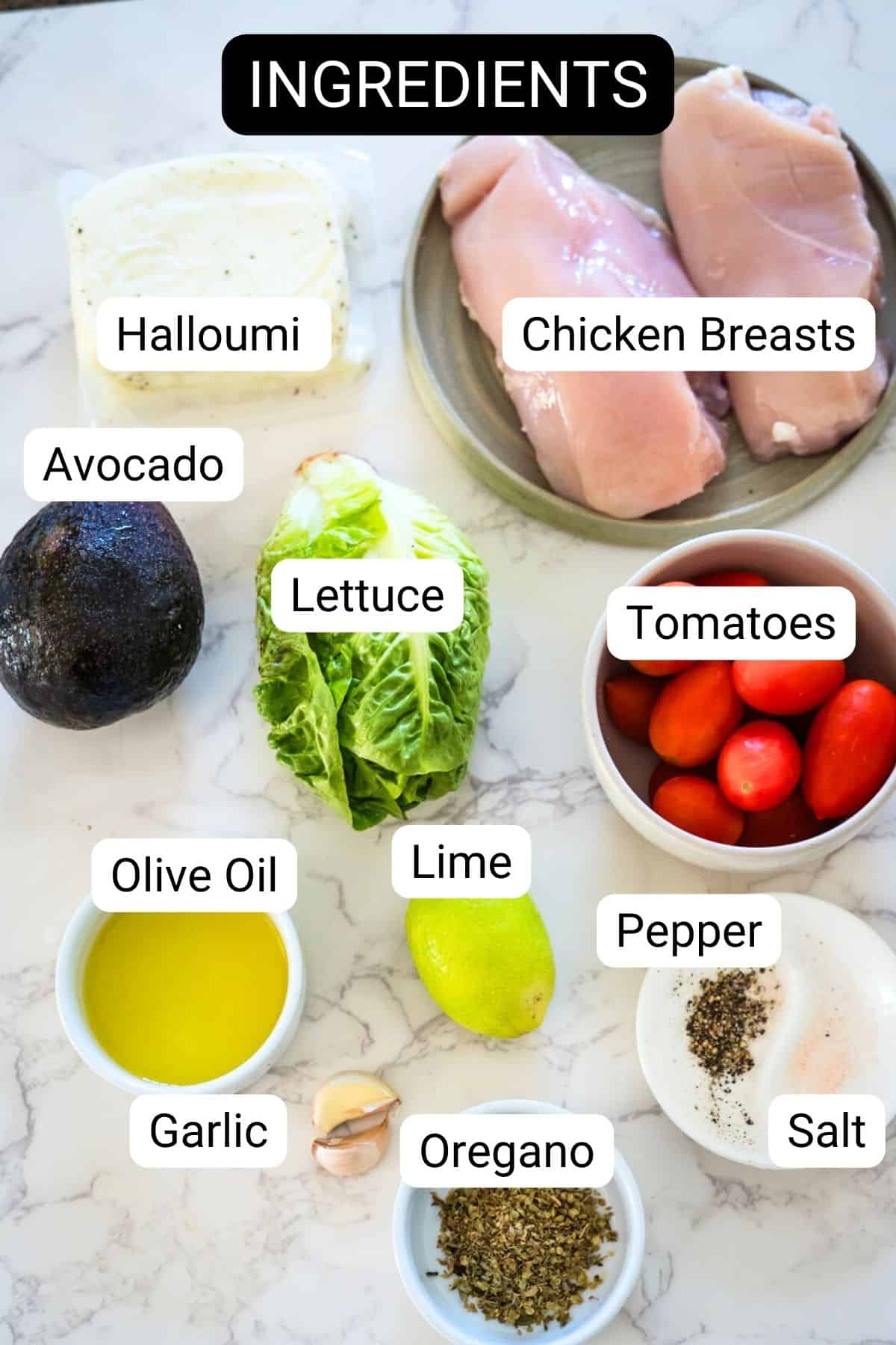 Ingredients for a recipe displayed on a marble countertop, labeled including halloumi, chicken breasts, avocado, lettuce, tomatoes, olive oil, lime, garlic, oregano, pepper, and salt.