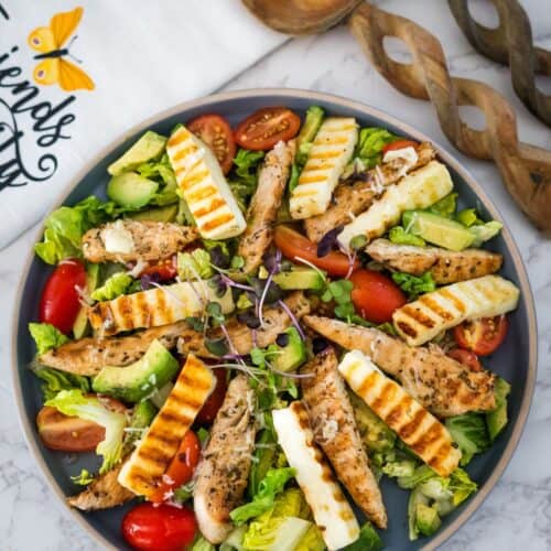 A plate of salad with grilled chicken, sliced avocado, tomatoes, and halloumi cheese, garnished with microgreens.