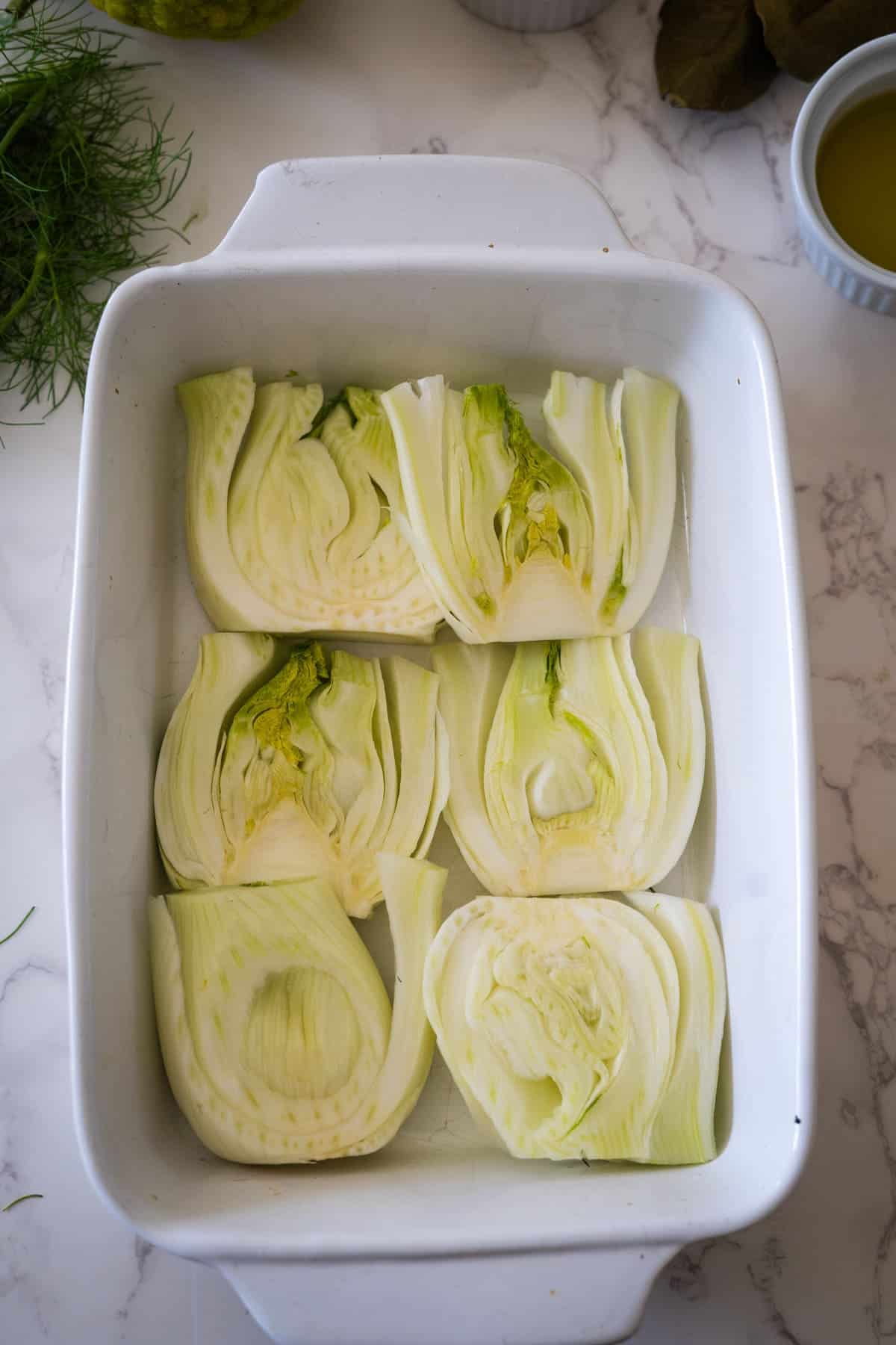 Halved fennel confit arranged in a white baking dish on a marble countertop.