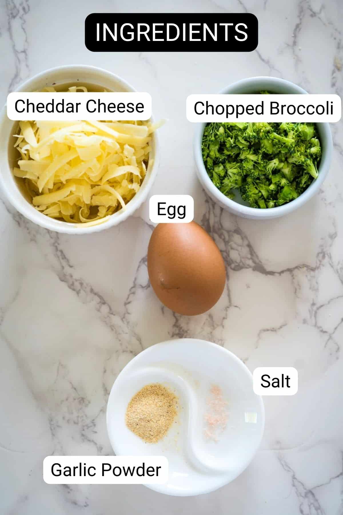 Ingredients for a broccoli cheddar waffles recipe: cheddar cheese, chopped broccoli, an egg, garlic powder, and salt, arranged on a marble surface.